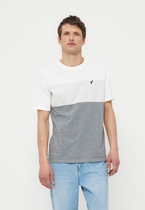 Άνδρας στέκεται σε λευκό φόντο φορώντας ένα μπλουζάκι με κοντά μανίκια σε λευκό, ανοιχτό γκρι και σκούρο γκρι με χρωματικούς μπλοκαρίσματα και ανοιχτό μπλε τζιν.