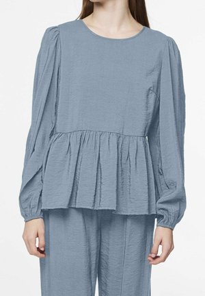 Vrouw draagt een lichtblauwe blouse met lange mouwen en een gestructureerde stof, voorzien van een geplooide peplum taille en bijpassende losse broek.