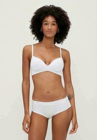 Bílý kompletní set spodního prádla, který zahrnuje bezešvé podprsenky s tenkými ramínky a designem ve tvaru písmene V, doplněný o odpovídající hipster kalhotky. Hladká textilie.