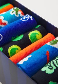 Chaussettes colorées présentées dans une boîte à motifs, présentant divers designs : bleu avec des nuages, fruits verts, rayures oranges et vagues noires.