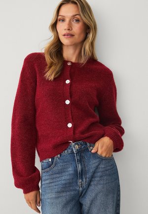 Woman with wavy blonde hair wearing a buttoned red cardigan and blue jeans, standing against a plain light gray wall.