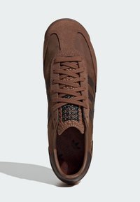 Brown athletic shoe with suede and leather materials, featuring three black stripes, textured accents, and flat laces.