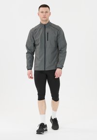 Grå idrottsjacka med hög krage, framficka med dragkedja och långa ärmar, kombinerad med svarta åtsittande leggings och idrottsskor.