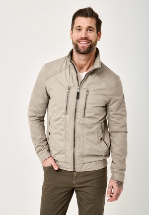 Lächelnder Mann mit Bart, der eine beige Jacke mit Reißverschluss und olivgrüne Hose trägt, steht mit einer Hand in der Tasche vor einem einfarbigen Hintergrund.