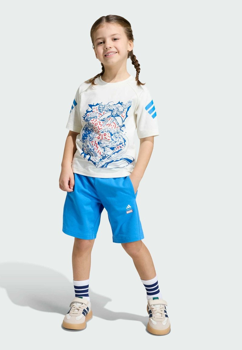 T-shirt blanc avec motif bleu, short bleu et baskets beige. Comprend des rayures bleues sur les manches et des chaussettes à rayures avec un accent vert.
