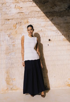 Ung kvinde iført en ærmeløs hvid striktop og lang sort nederdel, der står op ad en solbelyst tekstureret stenvæg.