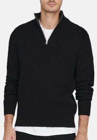 Schwarzer Zip-Hals-Pullover aus geripptem Strickstoff. Verfügt über lange Ärmel und einen weißen Innenkragen. Reißverschlussverschluss am Hals.
