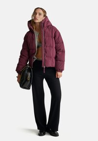 Burgundowa kurtka puchowa z wysokim kołnierzem, szarym wykończeniem wewnętrznym, czarnymi spodniami z szerokimi nogawkami oraz czarną torebką. Metaliczne akcenty na torebce.