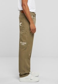 Pantalons cargo kaki avec poches latérales, marque KANI, et une étiquette en cuir à l'arrière. Portés avec des baskets blanches ornées de logo.