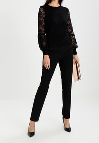 Haut noir avec manches en dentelle, col haut, associé à un pantalon noir ajusté. Tenant une pochette métallique rose doré. Talons pointus noirs.