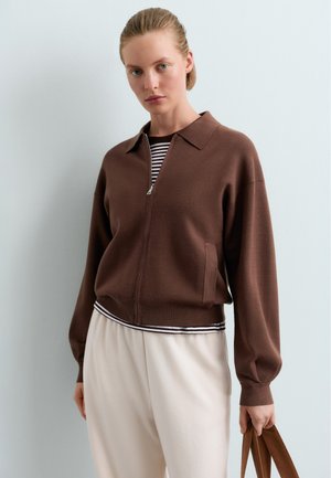 Žena nosi hnedý sveter na zips cez pruhované tričko a svetlé nohavice, drží hnedú tašku na jednofarebnom pozadí.