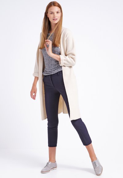 Un long cardigan beige sur un haut texturé gris et un pantalon marine. Le mannequin porte des chaussures grises à lacets et pose avec une main levée.