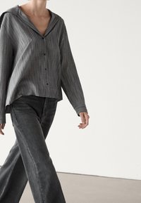Persona che cammina indossando una camicia grigia a righe larga con colletto ampio e jeans larghi grigio scuro su uno sfondo neutro.