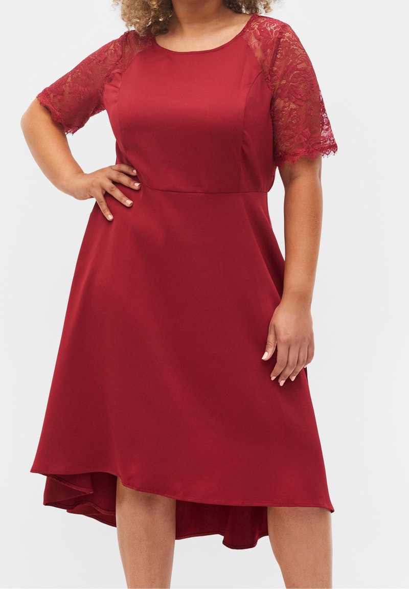 Femme portant une robe rouge mi-longue avec des manches en dentelle jusqu'aux coudes et un ourlet asymétrique, debout avec une main sur la hanche.