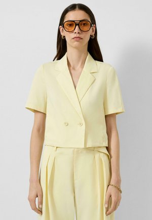 Frau trägt einen blassgelben Kurzarmblazer und passende plissierte Hosen, mit goldenen Creolen, Armband und bernsteinfarben getönten Sonnenbrillen.