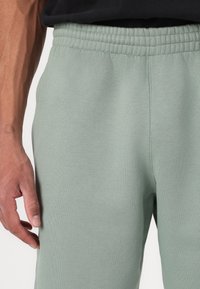 Ανοιχτοί πράσινοι φόρμες με λεία υφή, ελαστική μέση και στενή γραμμή. Κοντινή λήψη με το χέρι τοποθετημένο στο ύφασμα.