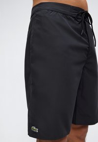 Μαύρα μαγιό βερμούδα με κορδόνι στη μέση και πράσινο λογότυπο κροκόδειλου κοντά στο αριστερό στρίφωμα, φορεμένα από ένα άτομο.