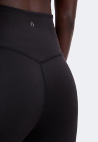 Gros plan d'une personne portant un legging noir taille haute avec des coutures visibles et un petit logo au centre du dos de la ceinture.