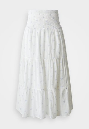 Valge kihtidega seelik, millel on smockitud vöökoht ja helebeežid dekoratiivmustrid. Valmistatud pehmest ja kergest kangast.