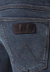 Džínové kalhoty s tmavě modrým nádechem, s koženou nášivkou s vyraženým nápisem "Wrangler" a charakteristickým cik-cak švem na zadní kapse.