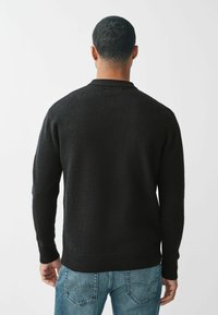 Čierny pletený sveter s okrúhlym výstrihom, rebrovanými manžetami a lemom. Nosený s svetlomodrými džínami, ktoré ukazujú textúrovanú látku. Pohľad zo zadu.
