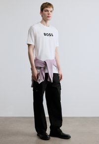 Valge t-särk musta "BOSS" logoga, kokku sobitatud lahtiste mustade cargo-pükstega ja helelilla kampsuniga, mis on vöökohale seotud. Mustad jalanõud.