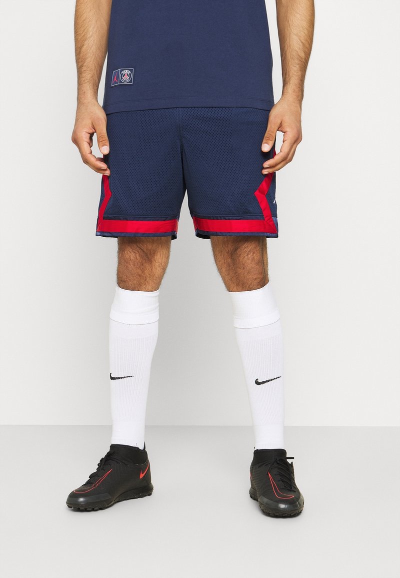 Homme portant un short de sport bleu marine avec des bordures rouges, des chaussettes blanches montantes jusqu'aux genoux avec un logo noir, et des crampons noirs avec des détails rouges.
