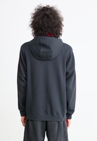 Sweatshirt preto com interior vermelho, apresentando um capuz com cordão, punhos e bainha canelados. As costas apresentam uma textura lisa sem logótipos.
