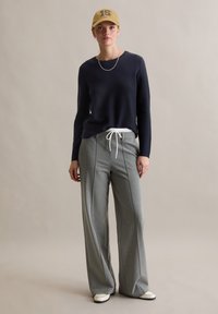 Marineblauer Pullover mit langen Ärmeln, kombiniert mit hellgrauen, weiten Hosen mit Seitennaht. Das Modell trägt eine beige Mütze mit einer Zahl.