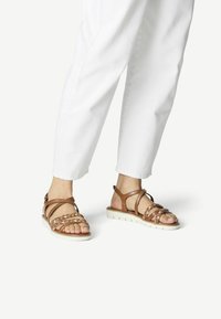 Sandalias de cuero marrón con correas trenzadas, una suela blanca acolchada y soporte para el tobillo, combinadas con pantalones blancos.