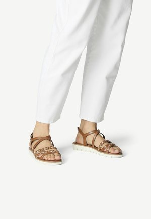 Sandales en cuir marron avec des brides tressées, une semelle blanche rembourrée et un soutien de cheville, assorties à un pantalon blanc.