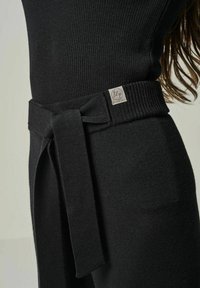 Schwarzes Strickkleid mit gerippter Textur, tailliertem Design und einem passenden Stoffgürtel mit Schleife sowie einem kleinen Logo-Detail am Bund.