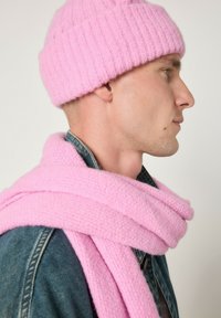 Rožinė megzta kepurė ir derantis šalikas, abu pagaminti iš minkštos, tekstūruotos medžiagos. Kepurė turi gofruotą dizainą; šalikas suvyniotas laisvai.