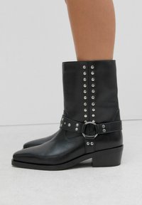 Botas de tobillo de cuero negro con punta afilada, tachuelas plateadas a lo largo del costado y una correa con hebilla, que cuentan con un tacón bajo en bloque.