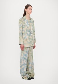 Light beige suit with blue and green palm leaf print. Features a single-button blazer and loose, wide-legged trousers. Black shoes.