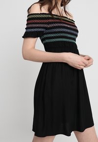 Musta olkaimeton mekko, jossa on monivärisiä brodeerattuja kuvioita, tiukka yläosa ja valuva hame lyhyillä hihoilla. Sileä kangas.