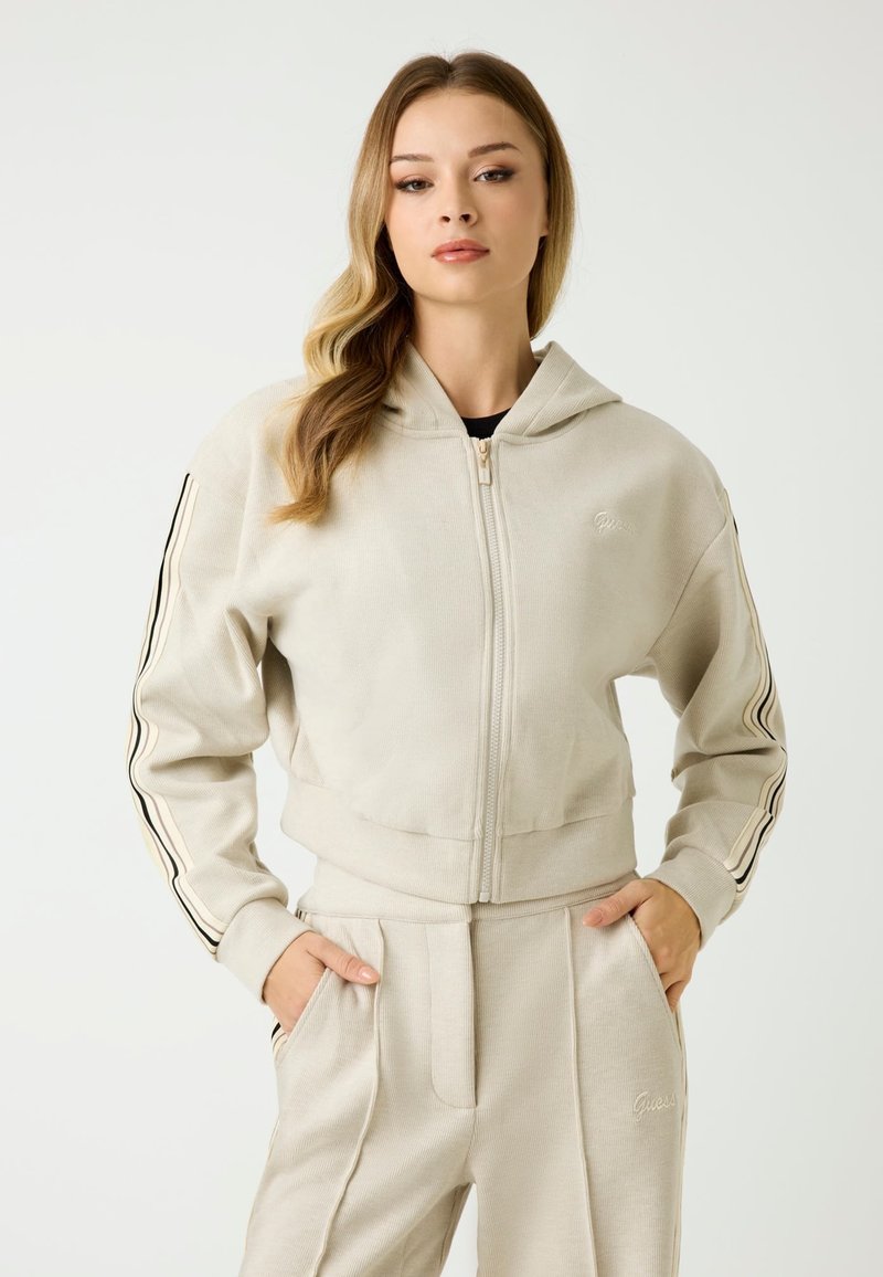 Beżowy krótki hoodie z zamkiem błyskawicznym z przodu i kapturem. Posiada dwie czarne paski na każdym rękawie oraz ribbrową teksturę. Haftowane logo na klatce piersiowej.