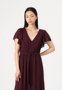 Burgundy dress with a V-neck, flutter sleeves, and a cinched waist. Made of lightweight fabric with a smooth texture and belted detail.