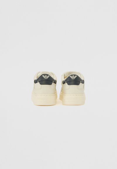 Sneakers with a cream-colored upper, navy leather accents, and textured rubber soles. Features a logo on the back and padded collar.