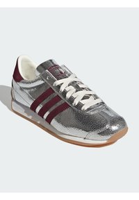 Hõbedased metallik sportlikud kingad burgundia triipudega, tekstureeritud pealisosa, valged paelaotsad ja kummikreen. Klassikaline disain, millel on ümar varvas.