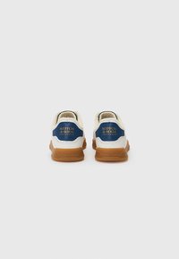 White sneakers with a blue accent, featuring a textured cream upper, round toe, and a gum rubber sole. "SCOTCH & SODA" logo on the heel.