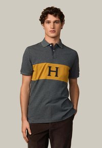 Šedé polo tričko s žltým vodorovným pruhom a námorníckym logom "H". Krátke rukávy, tradičný golier a zmes bavlny.