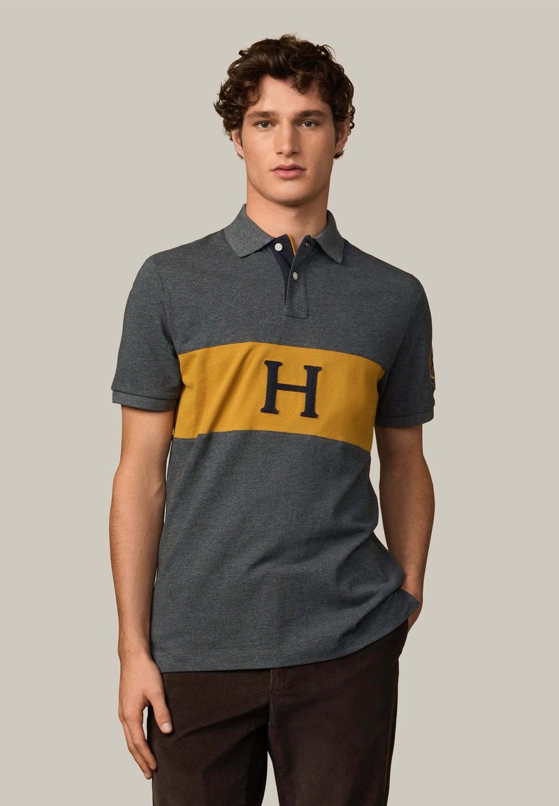 Šedé polo tričko s žltým vodorovným pruhom a námorníckym logom "H". Krátke rukávy, tradičný golier a zmes bavlny.