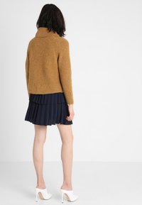 Brun stickad tröja med hög krage, kombinerad med en marinblå plissékjol och vita högklackade skor, sedd bakifrån.