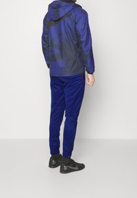 Dunkelblaue Sportjacke mit Kapuze und schwarzen Akzenten, kombiniert mit passenden blauen Jogginghosen und schwarzen Sportschuhen mit silbernen Details.