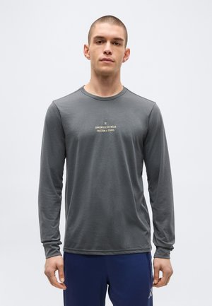 Muž sediašý v šedom tričku s dlhým rukávom s nápisom „Niekde medzi slobodou a chaosom“, stojaci pred jednofarebným svetlým pozadím.