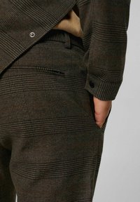 Gros plan d'une personne portant un costume à carreaux marron avec la main dans la poche latérale et un bouton visible au poignet de la veste.