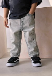 Jeans en denim gris légèrement usé avec une coupe décontractée, associés à des baskets à velcro noires avec des accents blancs. Tenue décontractée, adaptée à la vie quotidienne.
