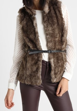 Vrouw met een bruine nep bontvest met een zwarte riem, een witte doorschijnende top met lange mouwen en donkerbruine leren broek, tegen een witte achtergrond.