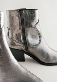 Bottines métalliques argentées avec une finition texturée, fermeture éclair sur le côté et un talon noir robuste. Présente des accents de couture minimalistes.